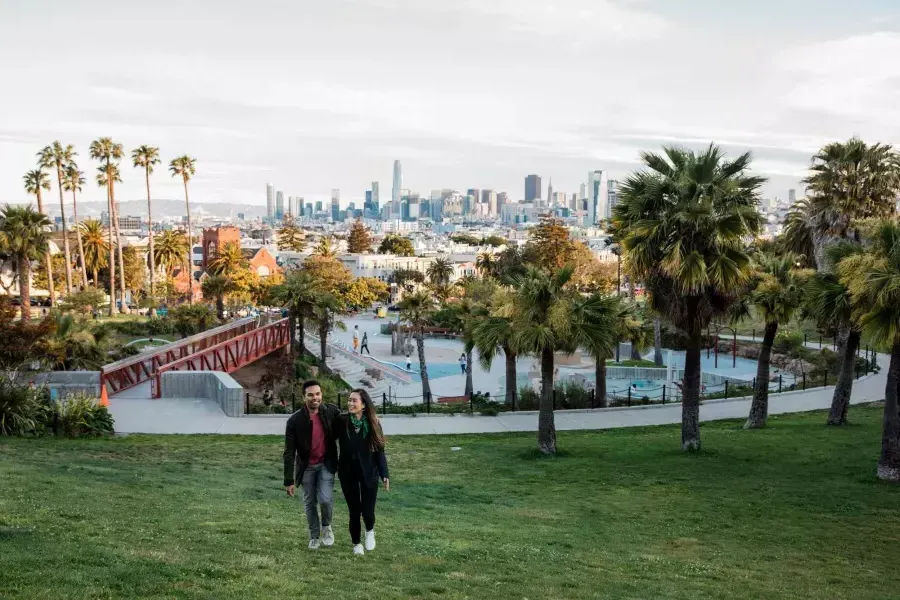 Un couple marche vers la caméra avec Dolores Park et la skyline de San Francisco derrière eux.